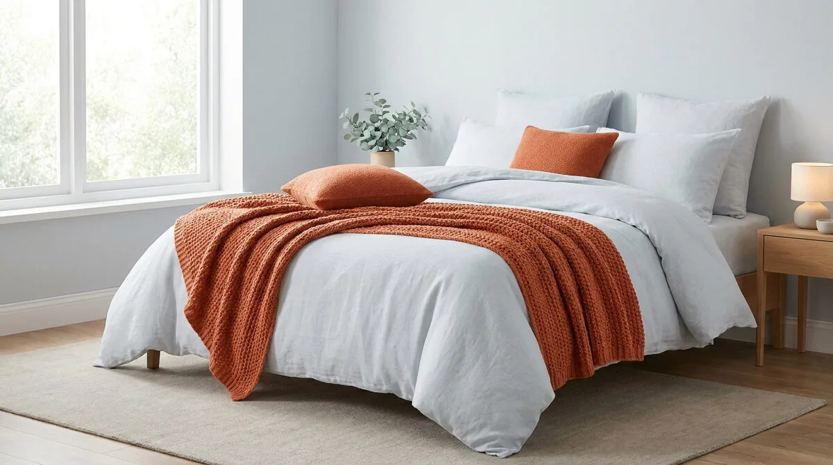 Lit avec linge de lit blanc, plaid en maille et coussins orange rouille dans une chambre lumineuse.