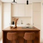 Petite cuisine lumineuse avec îlot central en bois, deux tabourets et rangements beiges minimalistes.