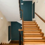 Escalier en bois moderne avec rampes noires, murs crème et mur d'accent bleu pétrole, éclairé par des rubans LED.