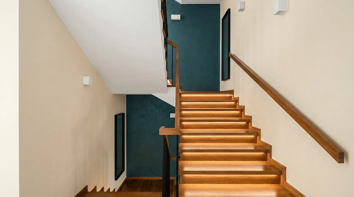 Escalier en bois moderne avec rampes noires, murs crème et mur d'accent bleu pétrole, éclairé par des rubans LED.