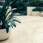 Mains avec gants de jardinage plaçant une plante dans un pot sur une terrasse carrelée moderne.